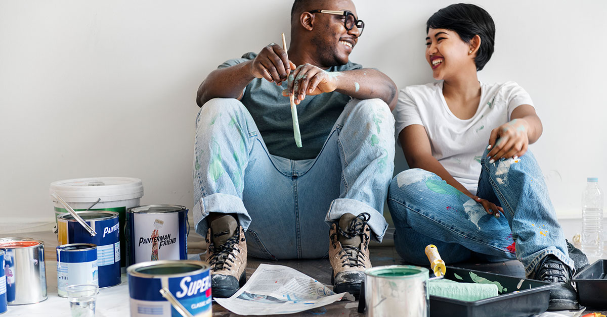 A couple sits on the floor surrounded by paint cans and painting supplies. They are covered in paint, looking at each other and smiling.