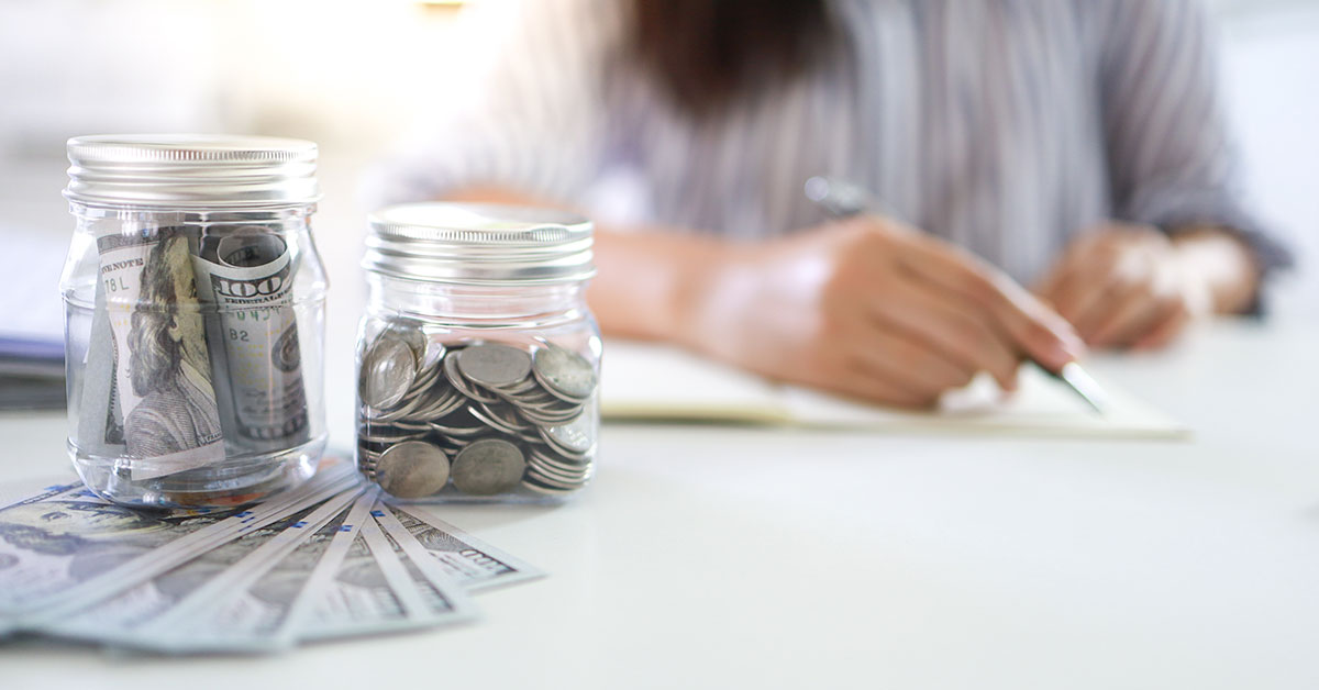 Close up of two jars full of coins and dollars sitting on top of a stack of $100 dollar bills. In the burred background is a woman writing at a table.