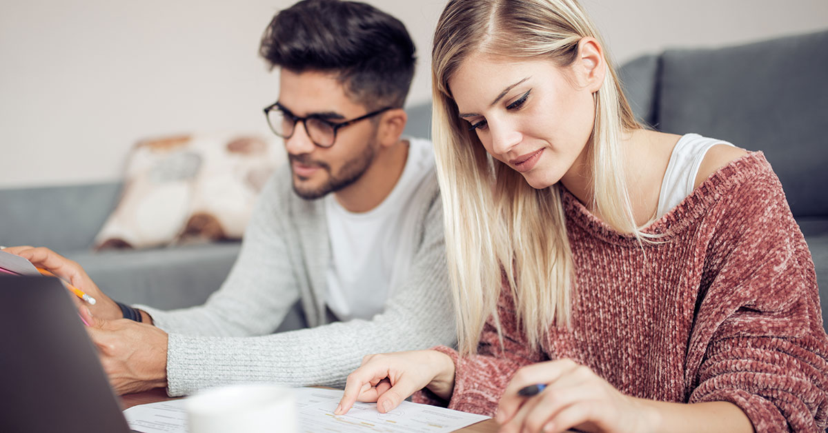 A man and woman sit at a coffee table together. The woman is using a calculator and the man is holding a paper and pencil