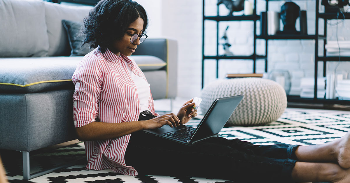 A young woman is sitting on the floor, leaning up against a couch. She has a laptop in her lap. She is typing and holding a credit card.