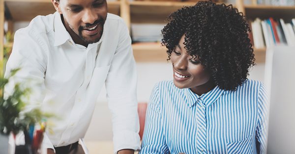 Woman in business attire sitting at a desk. A man stands next to her. They are both looking at some papers on the desk. The woman is pointing to the paper, and the man is writing