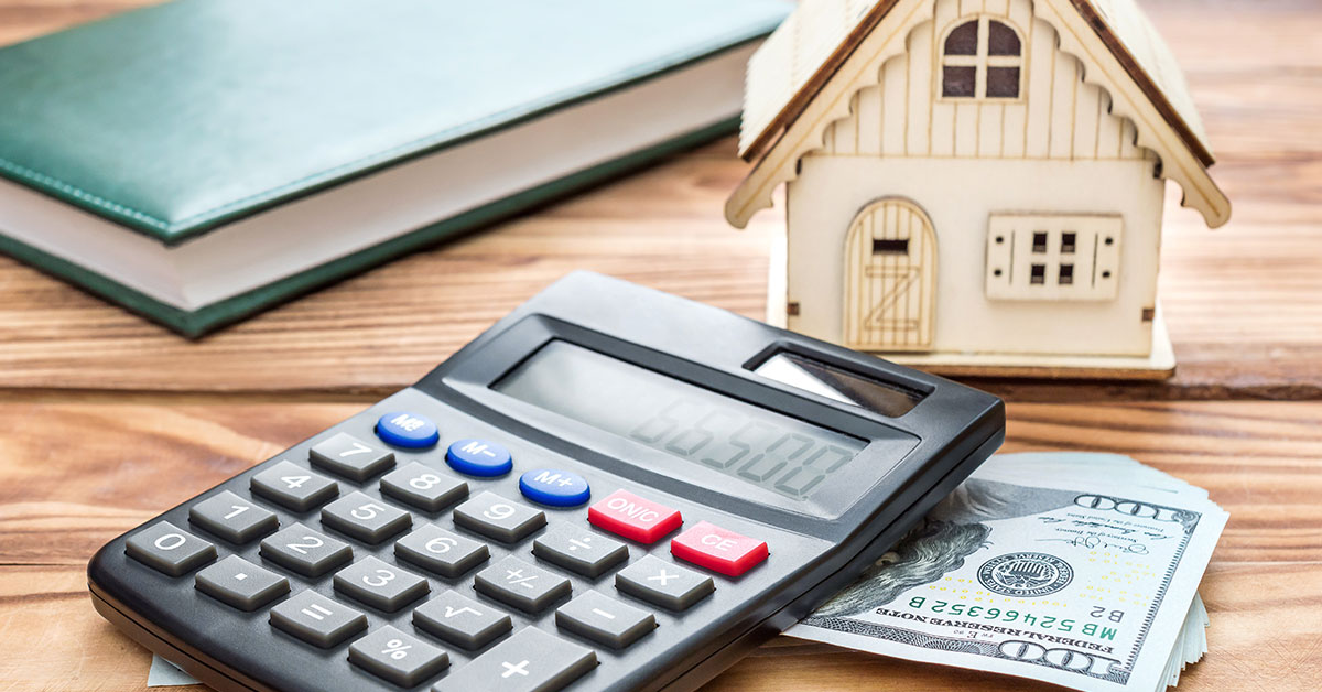 Photo of a calculator; stack of $100 US dollars; a green, leather journal; and a small wooden house figurine all resting on a wooden table.