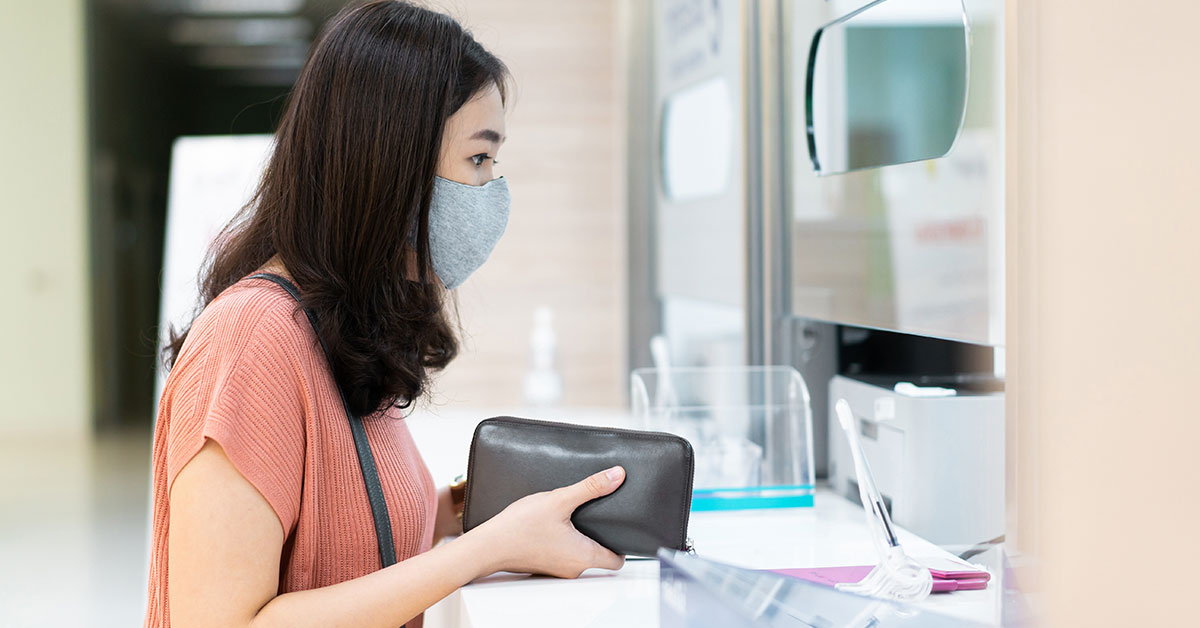 A woman is standing at a registration desk in a hospital, wearing a mask and holding her wallet.
