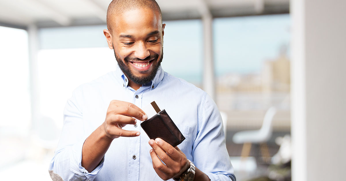 A man is standing alone in a room, smiling, pulling a credit card out of his wallet