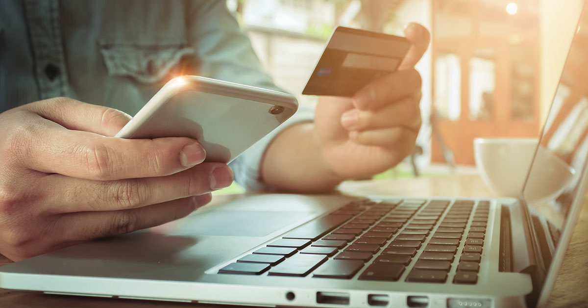 Close up of a man’s hands holding a cell phone and credit card. His hands are resting on a laptop.