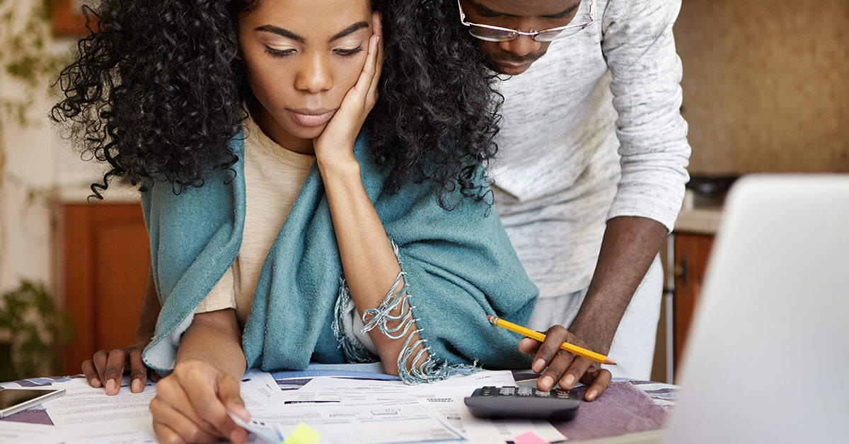 Photo of a young man and woman couple looking at a pile of bills scattered on a table. The woman is holding her head in her hand and sitting down. The man is standing behind her, leaning over and touching a calculator on the desk. They both have a neutral expression.