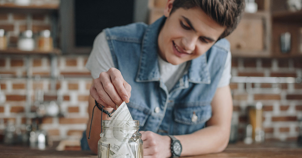 A teenage boy is standing over a table with a jar of money on it. He is smiling and reaching into the jar, pulling out a five-dollar bill.