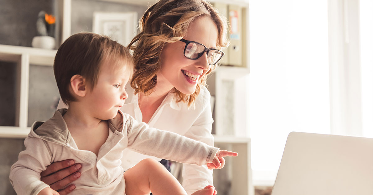 A woman is standing over a desk looking at her laptop and smiling. With one hand, she is typing, and with the other she is holding a toddler who is sitting on the desk.