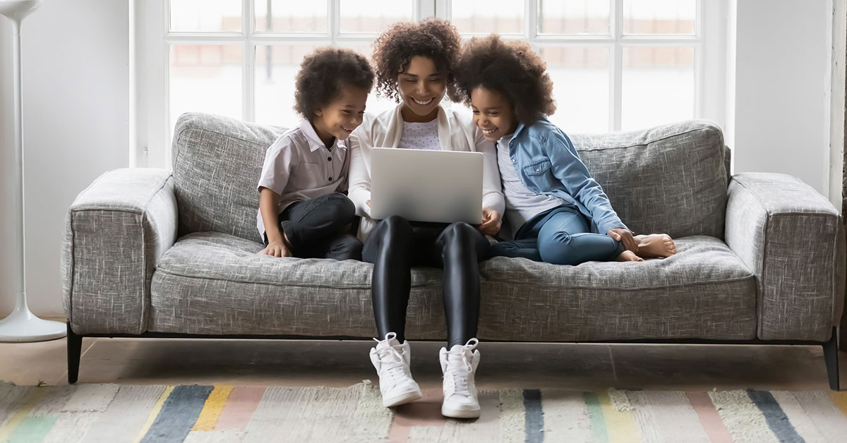 A woman sits on a couch with a laptop in her lap, smiling. Her young children are beside her.
