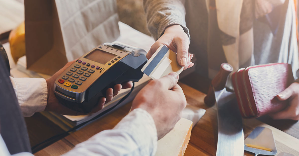 Close up of a person handing over a credit card to a clerk to pay for a purchase. The clerk is holding a payment processing machine.