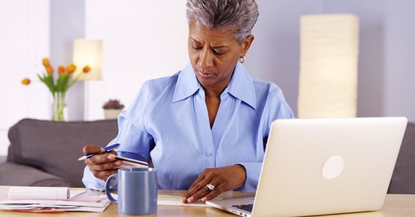 Older woman sitting at a table with computer, paperwork, and coffee mug. She is holding a pen and credit card.