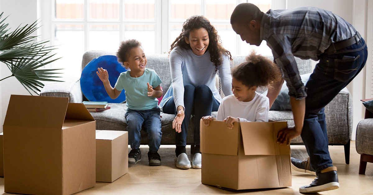 Family consisting of a mother, father, and two young children are playing in a living room. The mother and son sit on a couch, laughing and smiling. The daughter is inside an empty cardboard box and the father is playfully pushing her. The family is surrounded by empty cardboard boxes, indicating they just moved in.
