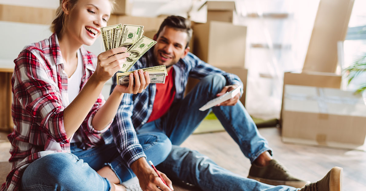 A man and woman couple sits on the floor among a stack of moving boxes. The woman is holding a stack of one-hundred-dollar bills and smiling.