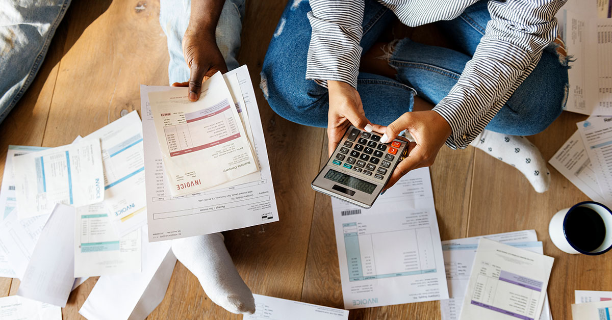 A couple sits on the floor with a pile of bills scattered around them. One person is holding a calculator and the other is holding some invoices.