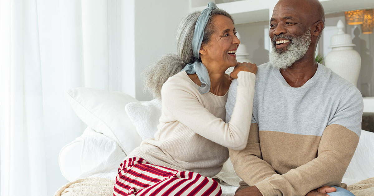 An older couple sits on a bed, smiling and leaning on each other.