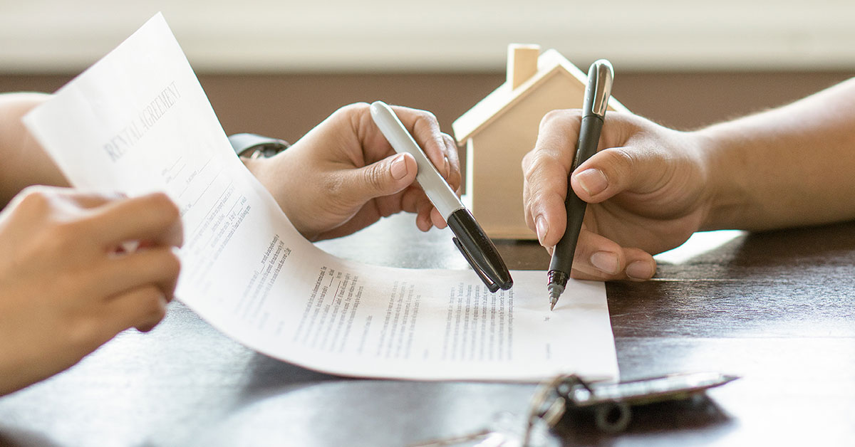 Photo close up of two people’s hands as they go over a contract on a table. The person on the left is holding the contract and pointing to the signature line. The person on the right is holding a pen, signing the contract. On the table is also a set of keys and a plastic house.