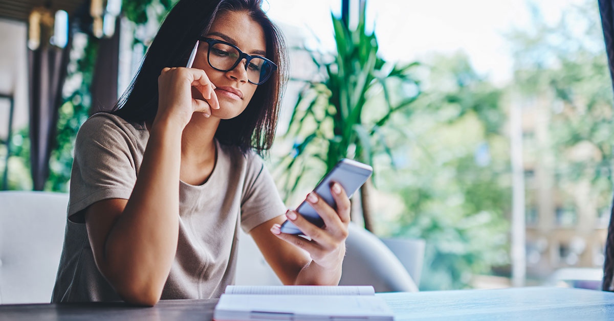 A teenage girl is sitting at a table. On the table is a book. She is looking down at her phone, which she is holding in her right hand. In her left hand, she holds a pencil.