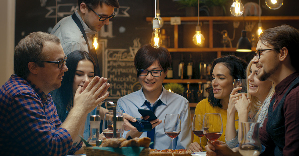 A group of thirty-something friends sit at a table in a restaurant having a conversation. The person in the middle is holding up their phone to a touchless pay machine being held by the waiter.