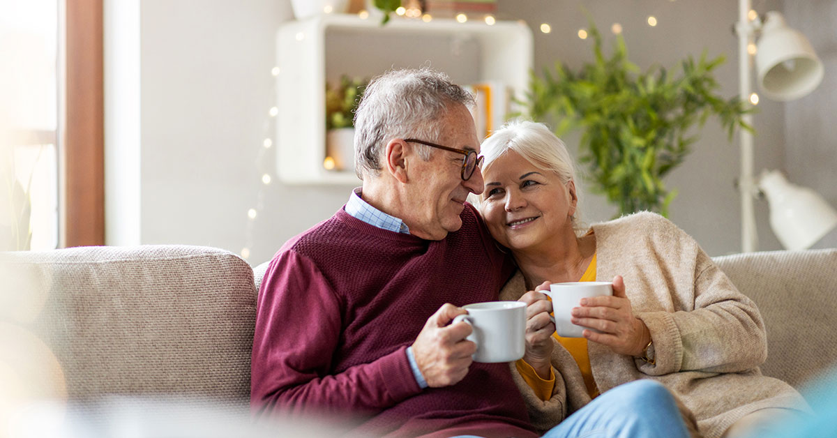 A senior couple sit together on the couch, holding coffee mugs, snuggled up and smiling at each other