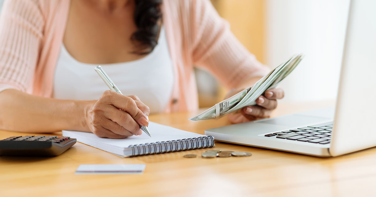 Close up of a woman’s hands as she sits at a desk holding dollar bills in one hand and writing in a notebook with the other. On the computer is an open laptop, calculator, some change, and a credit card.