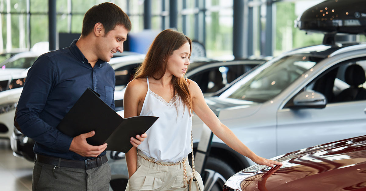 A woman touches a vehicle as she speaks with a car salesman about purchasing it.