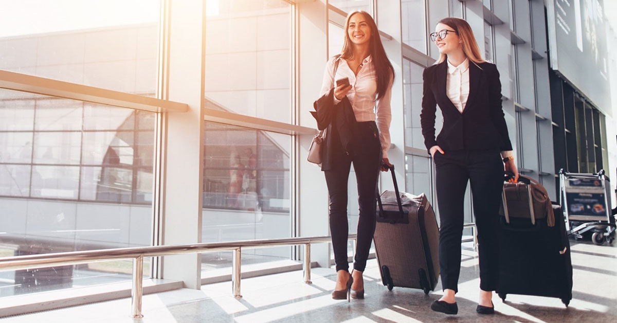 Two women professionals smile as they walk to their gate while traveling on a business trip.