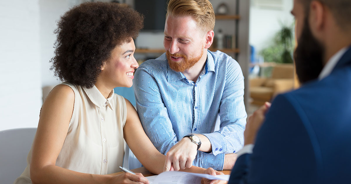 A smiling couple sits across from their lender at a desk as they sign their mortgage paperwork.