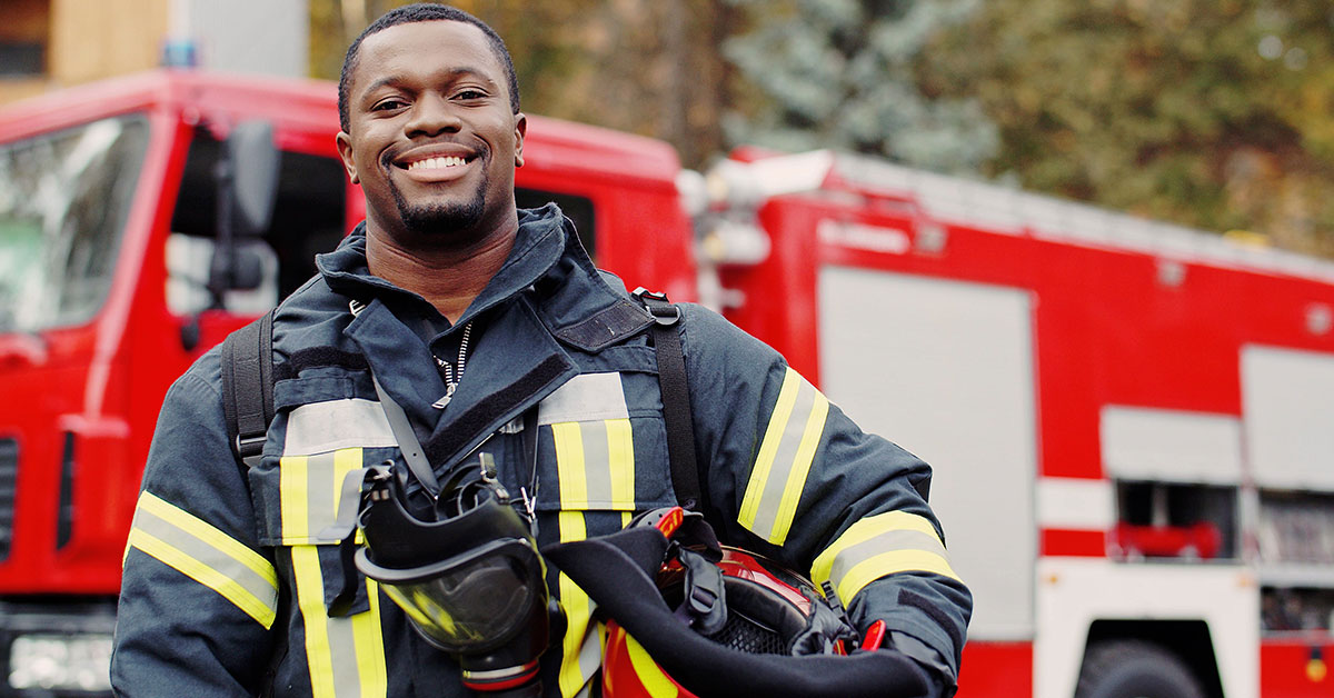 Fireman smiles at the camera holding his helmet in one arm with a firetruck in the background.