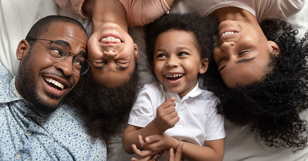 Family of four lays on a bed while smiling and laughing up at the camera. 