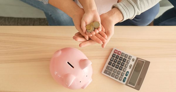 Top down view of the hands of a man, woman, and young child gathered together holding a few coins. Below their hands is a table with a pink piggy bank and a calculator