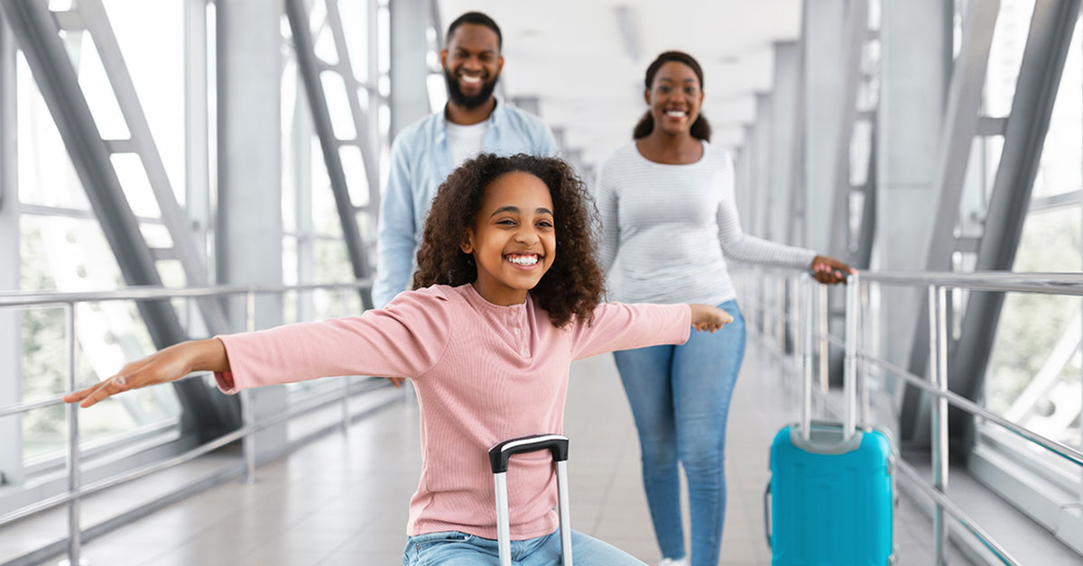 Couple walking through an airport terminal, with their daughter riding on a suitcase with her arms spread out to each side like an airplane.