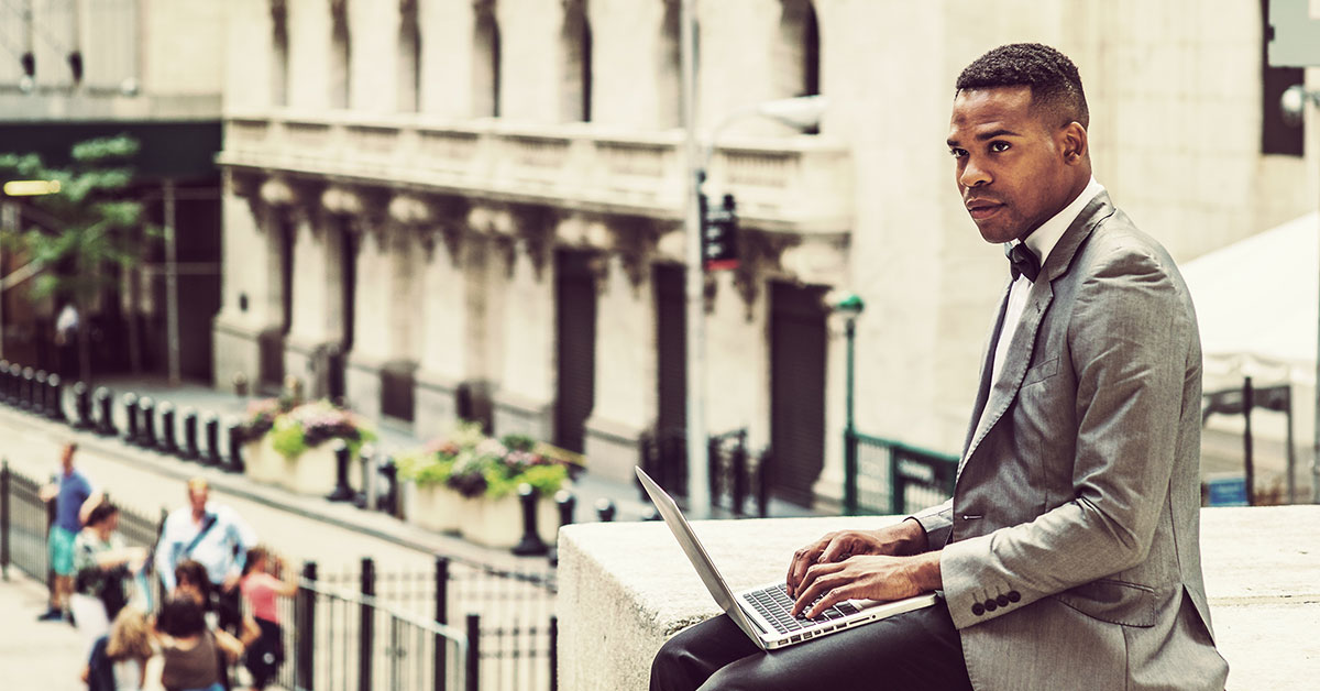 Man in business attire typing on a laptop, sitting on a concrete ledge. Below him, out of focus visitors can be seen looking at a historic building.