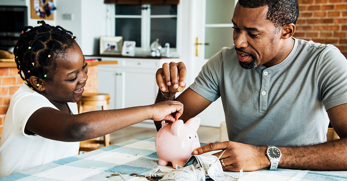 A father and his young daughter are seated at a kitchen table. On the table is a piggy bank surrounded by coins and dollar bills. Both the father and daughter are smiling and putting money in the piggy bank.