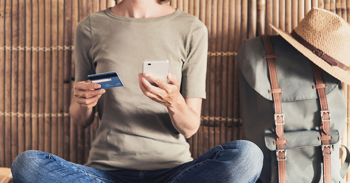 A woman is sitting on a bed. We cannot see her face, but we can see that she is holding a phone and credit card in her hands. Beside her on the bed is a backpack with a hat resting on top of it.