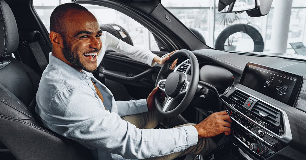 Inside the interior of a new car, a man sitting in the driver’s seat has a bright smile as he looks toward the viewer.