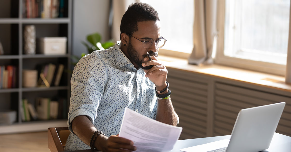 man sitting at a desk, focusing on his laptop while holding pieces of paper.