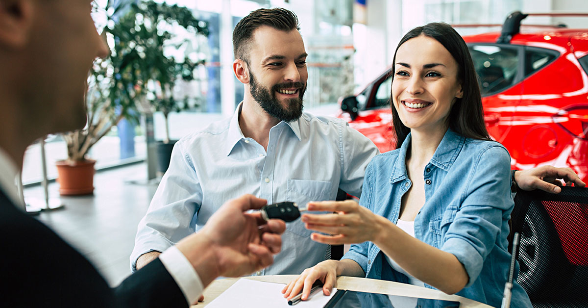 woman getting handed the keys to a car in the dealership, sitting next to a man.