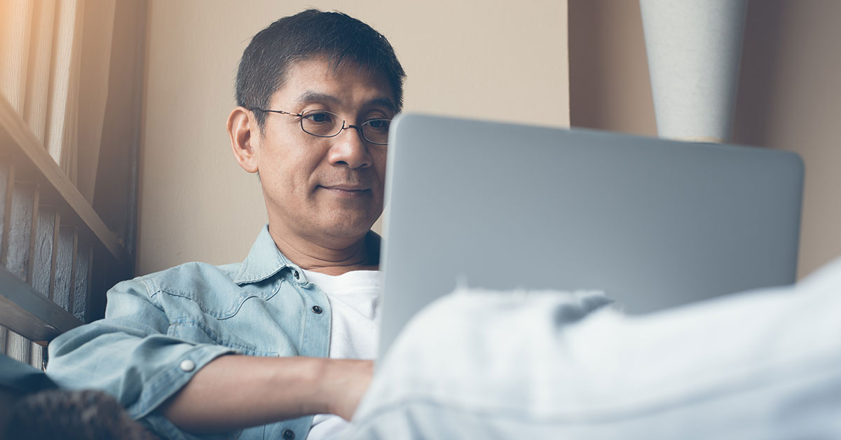 Middle-aged man sitting in a chair with a laptop resting on his lap. He is looking at the laptop screen and typing.