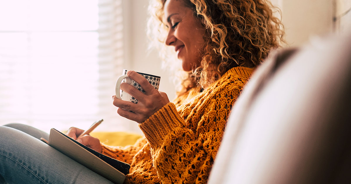 Side view of a middle-aged woman sitting on a couch. She is wearing a sweater and holding a mug in her hand. She is also writing in a notebook with a pen and smiling.