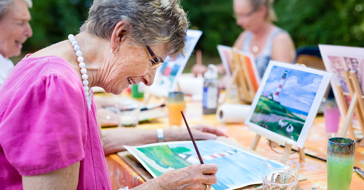 Elderly woman seated at table with friends, painting a lighthouse landscape