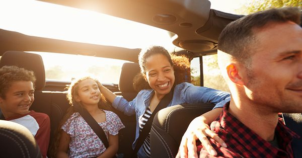 A close up view of a family in a car with the sunroof open. The sun is setting behind them. The father sits in the front seat and the mother is in the back seat with her two children, a son and daughter