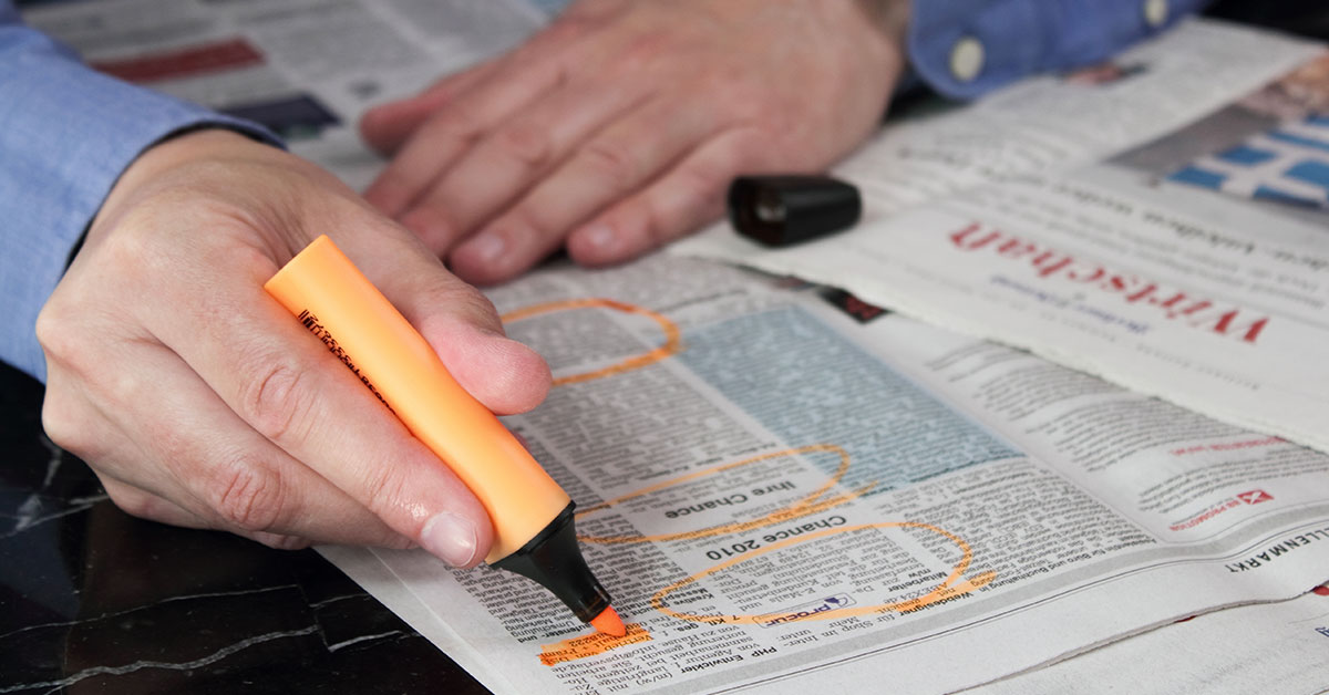 Close up of a man at a desk. We see that he is highlighting job ads in a newspaper.