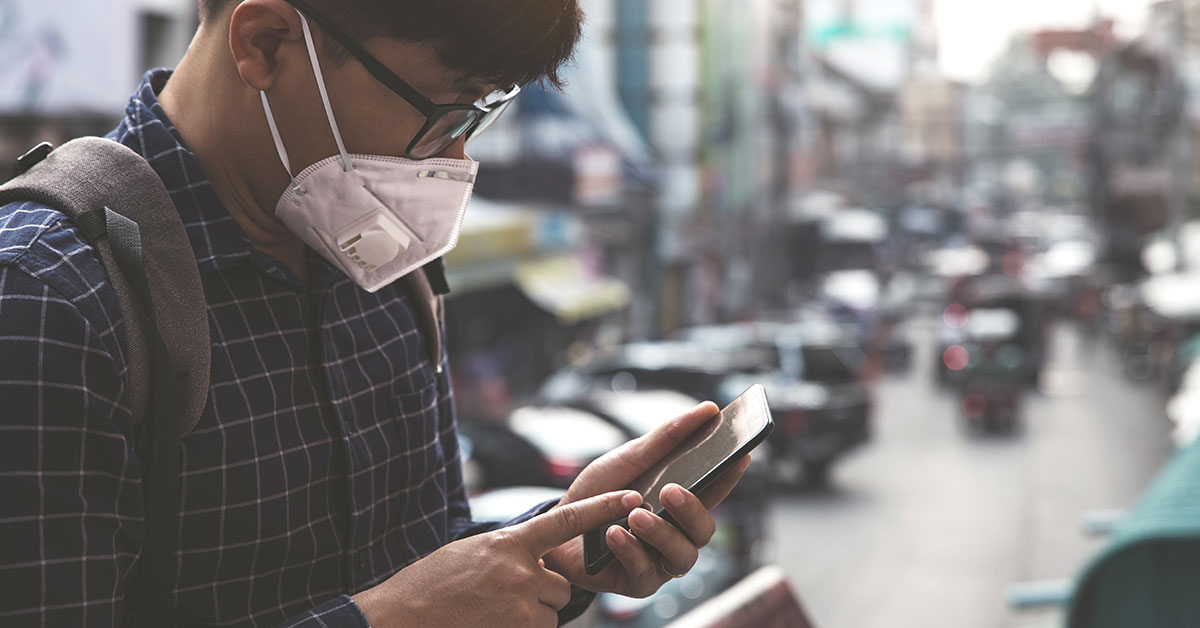 A young man wearing an N95 mask is standing outside looking at his phone. In the out-of-focus background, we can see that he is standing in a city with several cars and buildings behind him.