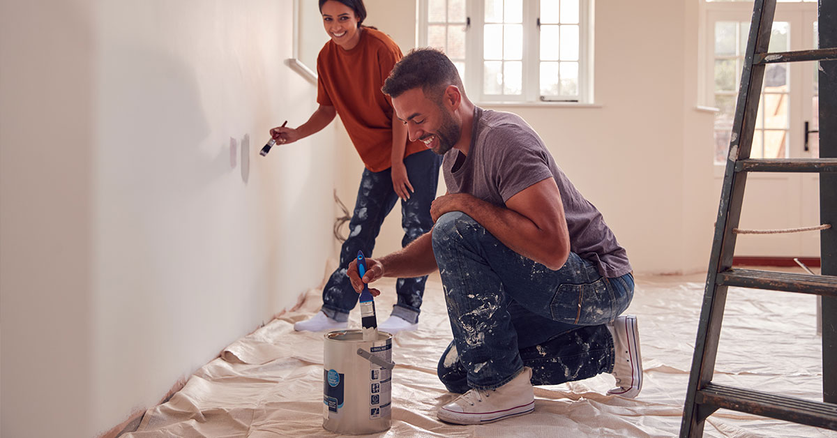 Smiling man is kneeling down to dip his paintbrush in the paint bucket as his partner smiles and paints the wall in the background