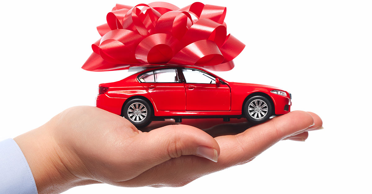 Up close photo a woman’s hand holding a red toy car. On top of the car is a very large red bow.