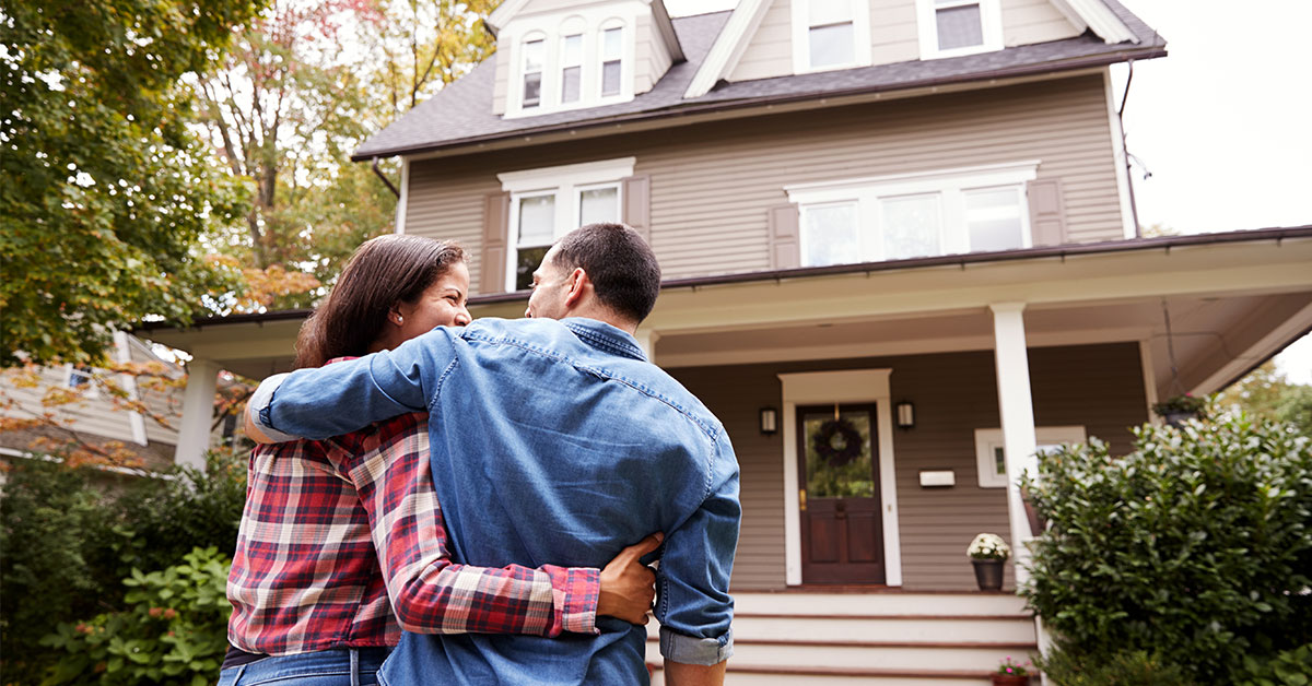 Couple smiles at each other with arms wrapped around one another as they walk towards their new home.