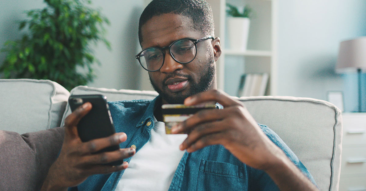 A man is sitting on a couch looking at his cell phone, which he is holding in his right hand. In his left hand, he is holding a credit card.