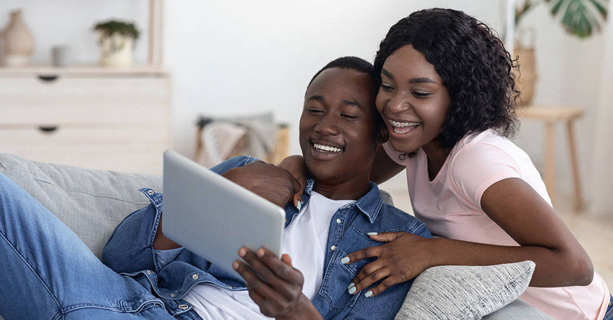 Young adult couple seated around a couch in their living room, smiling while looking at their online banking accounts on a tablet computer.