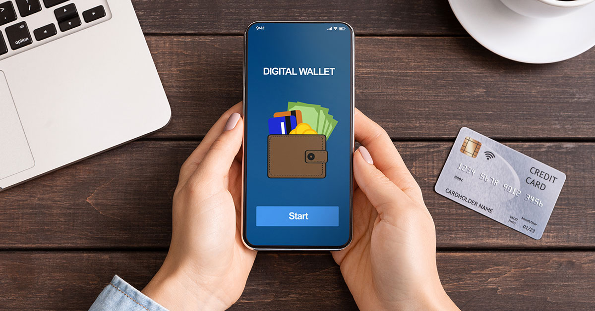 A woman’s hands are holding a phone. On the phone are the words “digital wallet” alongside a graphic icon of a wallet and a button that says “start.” Her hands are resting on a table next to a credit card, laptop, and cup of coffee.