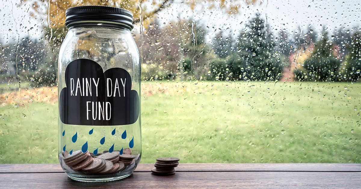 Clear glass jar labeled “Rainy Day Fund” with coins in it sits on a table in front of a rainy window.